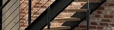 Ein Treppengeländer aus schwarzem Metall mit Holzstufen. Es befindet sich an einer roten Ziegelwand.
